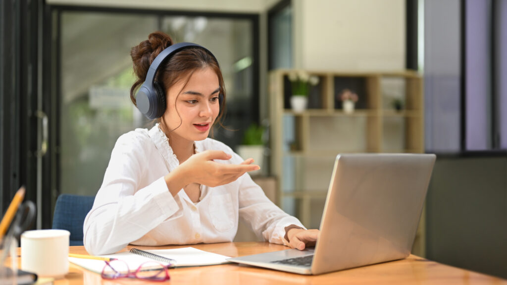 Young female employee wearing headphone and communicating by video conference on laptop computer.